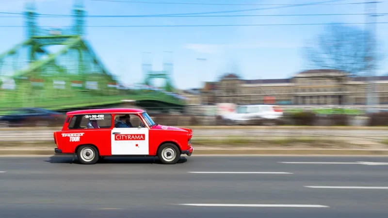 Piros-fehér trabant túraautó mozgásban a budapesti rakparton, elmosódott Szabadság híddal a háttérben.