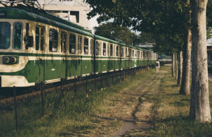 Zöld HÉV vonat mellett nő sétál egy fás, napsütötte ösvényen Budapesten, a MÜPA közelében.