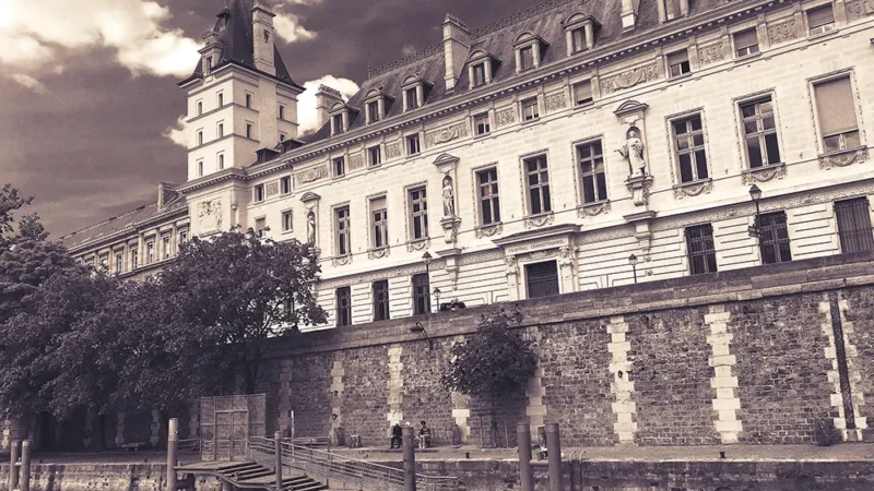 A párizsi Igazságügyi Palota sepia tónusú felvétele a Szajna partjáról, egy kis kikötővel előtérben.