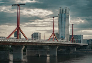 A budapesti Rákóczi híd vörös pilonjaival, a háttérben a MOL toronnyal és drámai, felhős égbolttal.